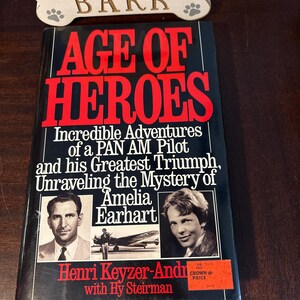 May include: A hardcover book titled "AGE OF HEROES" with red and white lettering. The book cover features images of Amelia Earhart and a man, with text detailing the book's subject matter. The book is set against a dark background.