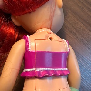 May include: A doll with red hair and a pink top with white trim. The doll's back is visible, showing a battery compartment and the word "Disney" imprinted on the neck. The doll's skin is a light peach colour.