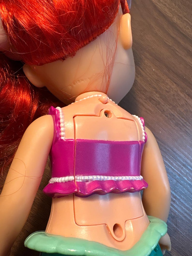 May include: A doll with red hair and a pink top with white trim. The doll's back is visible, showing a battery compartment and the word "Disney" imprinted on the neck. The doll's skin is a light peach colour.