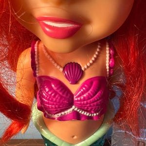 May include: A close-up of a doll with a mermaid design. The doll has bright red hair, a pink seashell bra, and a pearl necklace. The mermaid's tail is teal and green.