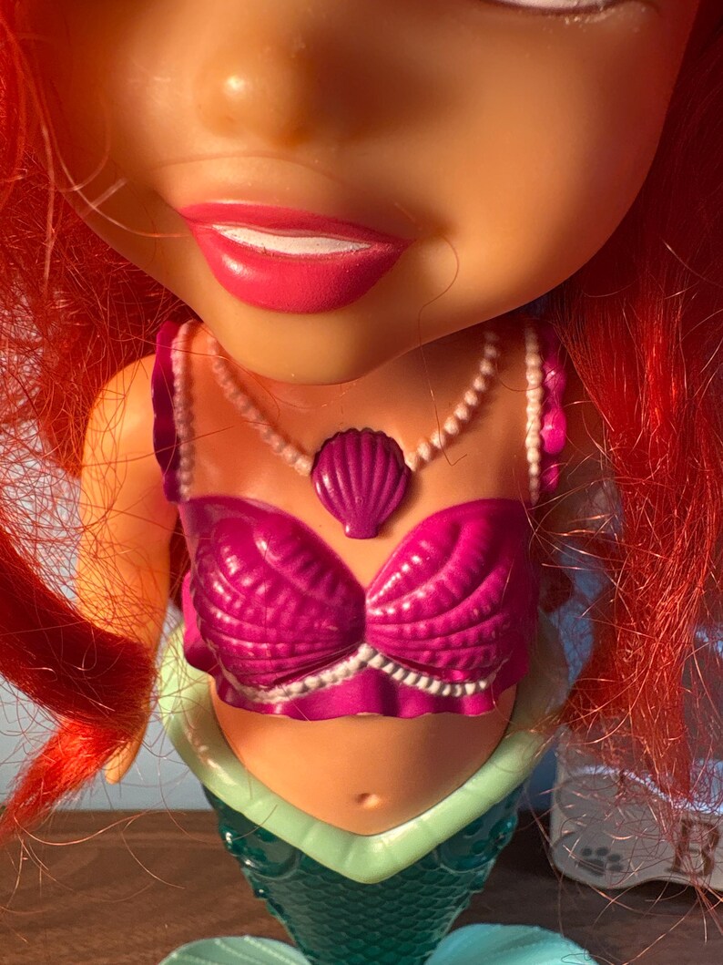 May include: A close-up of a doll with a mermaid design. The doll has bright red hair, a pink seashell bra, and a pearl necklace. The mermaid's tail is teal and green.