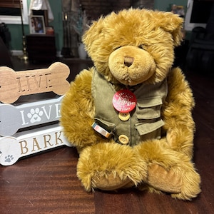 May include: A plush brown teddy bear wearing a green vest with buttons and a red tag. The bear is sitting in front of wooden dog bone-shaped signs that read "LIVE", "LOVE", and "BARK".