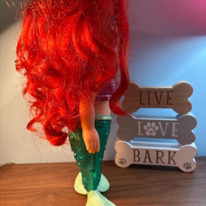 May include: A mermaid doll with bright red hair, a purple top, and a green tail. The doll is next to a wooden sign that says "LIVE LOVE BARK".
