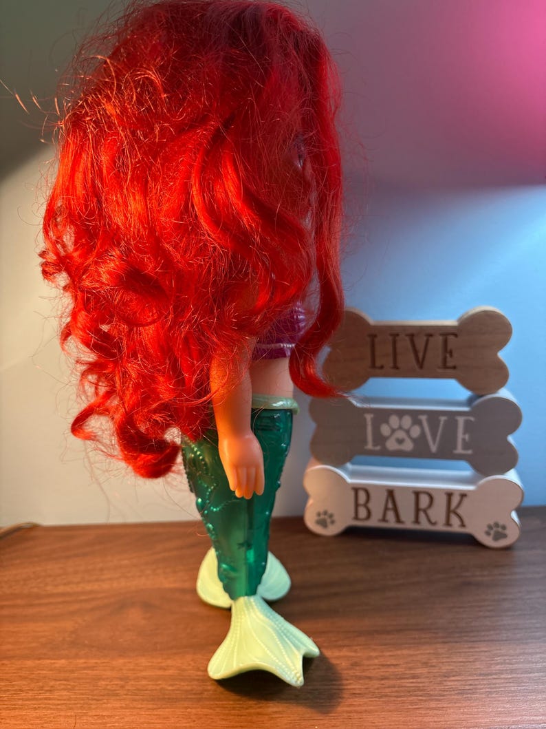 May include: A mermaid doll with bright red hair, a purple top, and a green tail. The doll is next to a wooden sign that says "LIVE LOVE BARK".