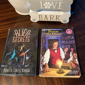 May include: Two paperback books, one titled "Alien Secrets" with a dark blue cover and a black and white illustration of a woman and a creature, and the other titled "The Story of Thomas Alva Edison" with a yellow cover and a color illustration of a man in a red vest and a white shirt standing in a room with a light bulb.