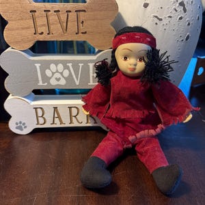 May include: A red and black doll with a beaded headband and black hair. The doll is sitting in front of three wooden bone-shaped signs that read "LIVE", "LOVE", and "BARK".