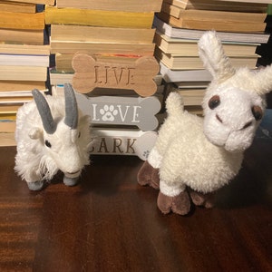 May include: Two plush toys, a white goat and a white llama, sit on a wooden surface in front of three wooden bone-shaped signs that read "Live", "Love", and "Bark".
