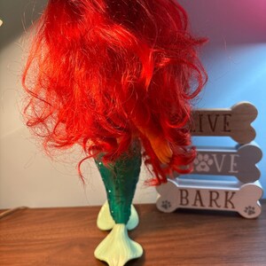 May include: A mermaid figurine with bright red hair and a green tail. The tail has a textured, scale-like pattern and fades into a light green fin. The figurine is placed in front of a decorative dog bone sign.