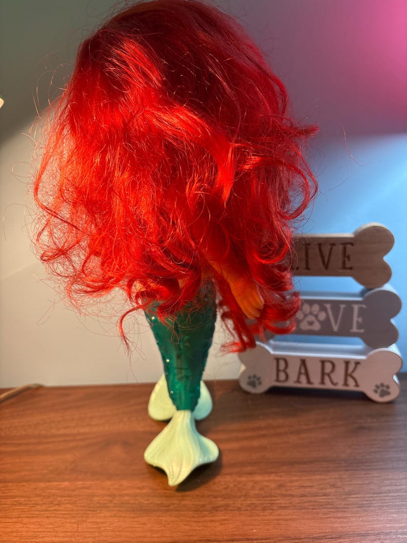 May include: A mermaid figurine with bright red hair and a green tail. The tail has a textured, scale-like pattern and fades into a light green fin. The figurine is placed in front of a decorative dog bone sign.