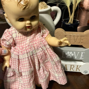 May include: A vintage porcelain doll wearing a pink and white gingham dress. The doll is standing next to a wooden sign that says "Live, Love, Bark".