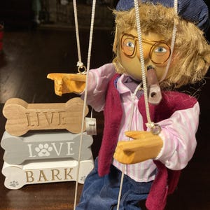 May include: A wooden puppet with a red sweater, blue pants, and a blue hat. The puppet is holding a pipe in its mouth and is wearing glasses. The puppet is hanging from strings and is in front of a wooden sign that says "Live Love Bark".