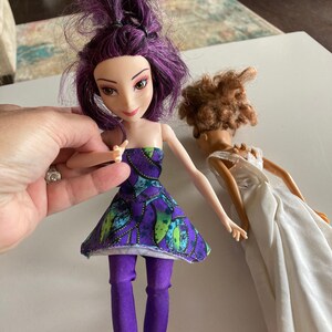 以下が含まれることがあります： 2体のドール。1体は紫色の髪に紫と緑の模様のドレスと紫色のレギンスを着用。もう1体は茶色の髪に白いドレスを着用している。