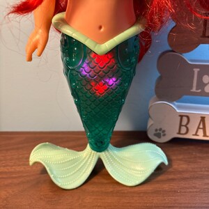 May include: A mermaid doll with red hair, a purple top, and a green tail. The tail has a scale pattern and a light green fin. The doll is on a brown surface.