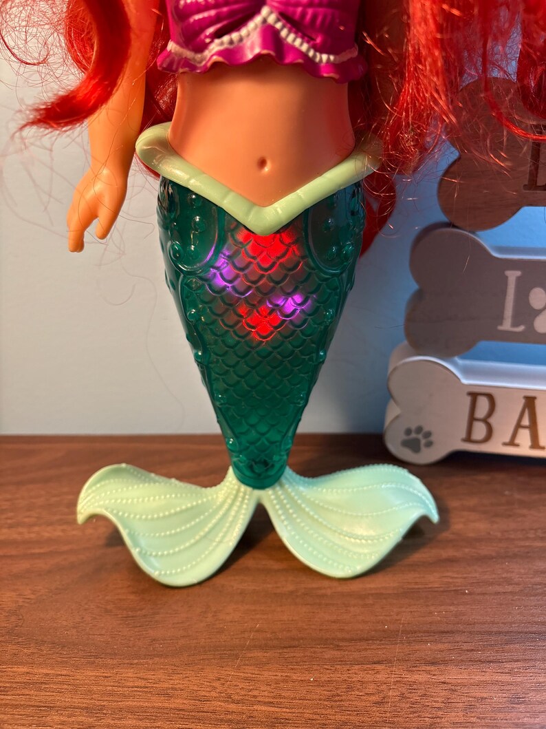 May include: A mermaid doll with red hair, a purple top, and a green tail. The tail has a scale pattern and a light green fin. The doll is on a brown surface.
