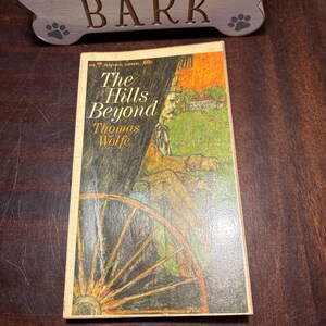 May include: A vintage paperback book titled "The Hills Beyond" by Thomas Wolfe. The cover features an illustration of a man in a carriage with a large wheel, set against a backdrop of rolling hills and an orange sky. The book is placed near a decorative dog bone-shaped sign that says "BARK".