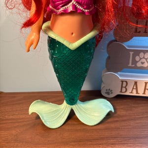 May include: A mermaid doll with red hair, a magenta seashell top, and a green tail. The tail has a scale pattern and light green fins. The doll is on a wooden surface.