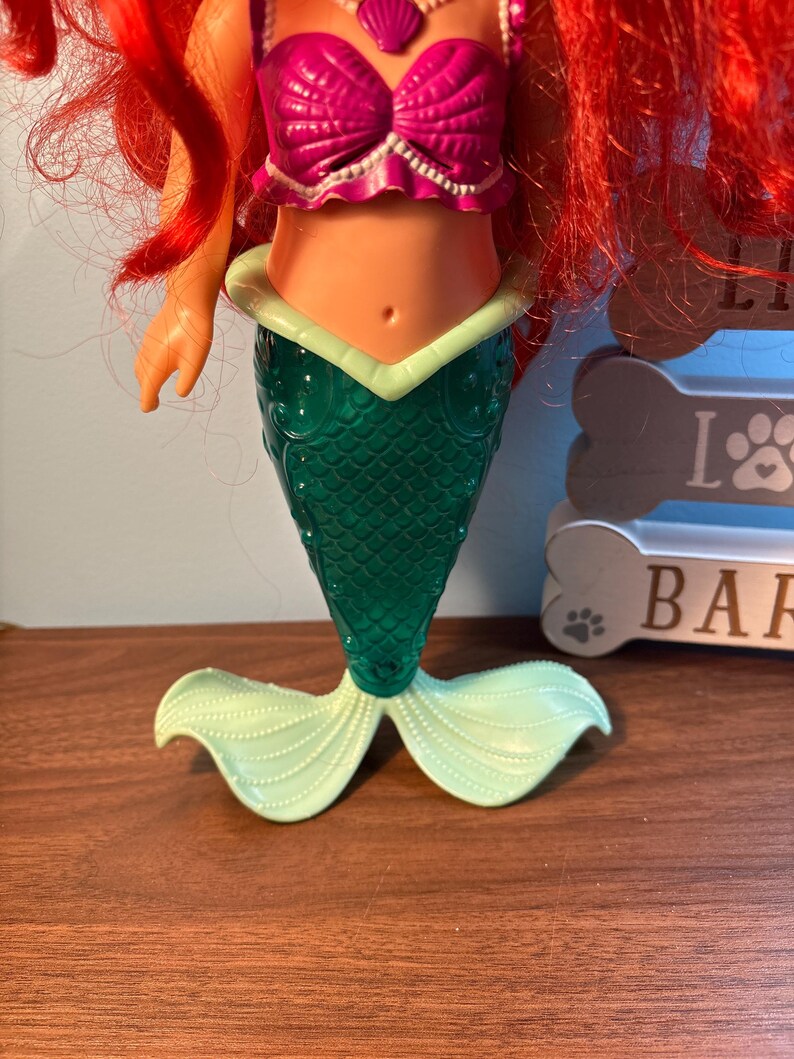 May include: A mermaid doll with red hair, a magenta seashell top, and a green tail. The tail has a scale pattern and light green fins. The doll is on a wooden surface.