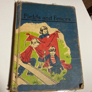 May include: Vintage book titled "Fields and Fences" with a blue cover and a colorful illustration. The illustration depicts three children with a red sheet and a wooden fence. The book's cover is worn, showing signs of age.