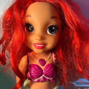 May include: A doll of a character with vibrant red hair, large blue eyes, and a pink seashell top. The doll has a fair complexion and a small, pink seashell pendant. The doll's lower body is green, resembling a mermaid's tail.