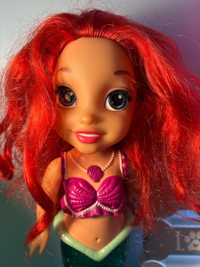 May include: A doll of a character with vibrant red hair, large blue eyes, and a pink seashell top. The doll has a fair complexion and a small, pink seashell pendant. The doll's lower body is green, resembling a mermaid's tail.