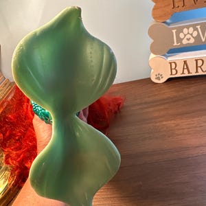 May include: A light green, abstract sculpture with a smooth, glossy finish. The sculpture has a unique, biomorphic shape, with a narrow waist connecting two larger, rounded forms. The background includes a wooden sign with the words "LIVE LOVE BAR" and a paw print.