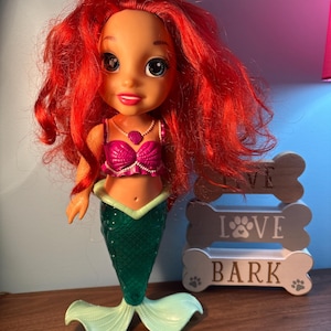 May include: A mermaid doll with vibrant red hair, a pink top, and a green tail. The doll wears a pearl necklace and stands beside a wooden sign that reads "LOVE BARK".