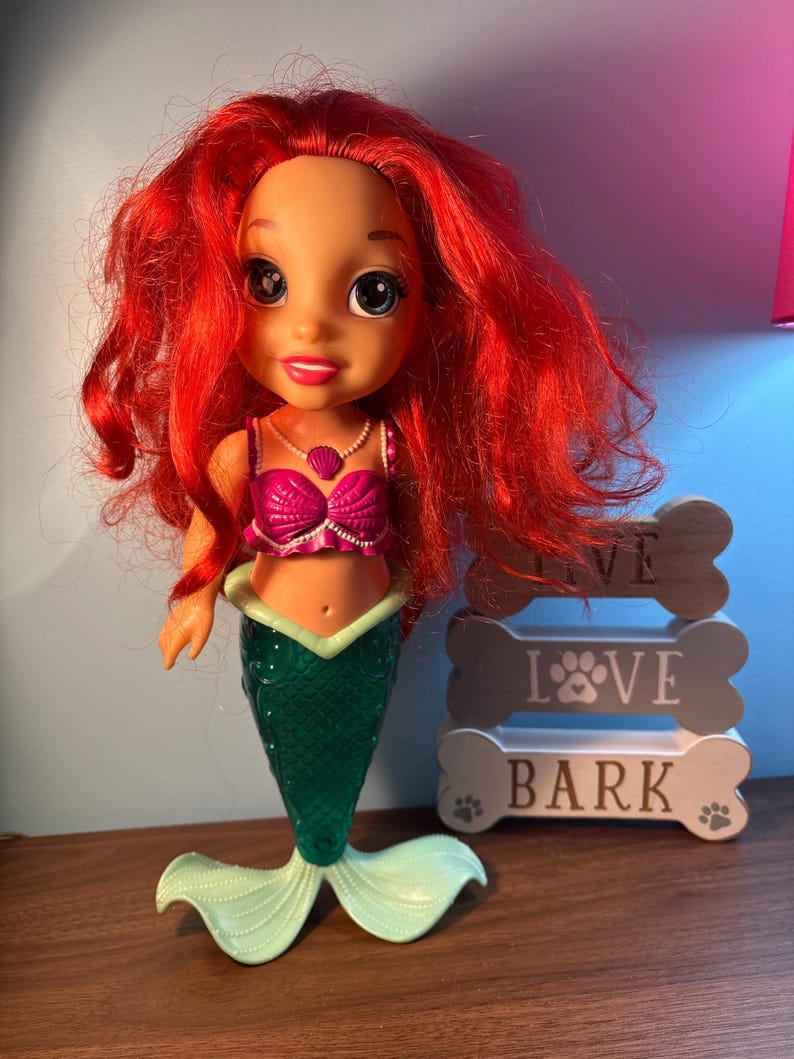 May include: A mermaid doll with vibrant red hair, a pink top, and a green tail. The doll wears a pearl necklace and stands beside a wooden sign that reads "LOVE BARK".