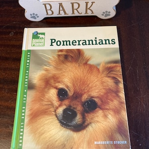 May include: A book titled "Pomeranians" from the Animal Planet Pet Care Library series. The cover features a close-up photo of a Pomeranian dog with a soft, fluffy, orange coat.