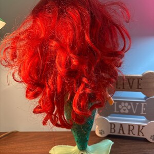 May include: A mermaid figurine with bright red hair and a green tail. The tail has a light green fin. The mermaid's hair is styled in loose curls. The figurine is a decorative item.