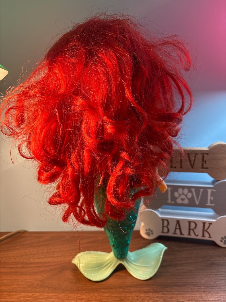 May include: A mermaid figurine with bright red hair and a green tail. The tail has a light green fin. The mermaid's hair is styled in loose curls. The figurine is a decorative item.
