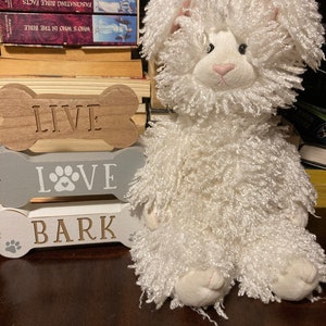 May include: A white fluffy stuffed animal shaped like a dog sitting on a wooden surface. The dog has pink ears and a pink nose. The dog is sitting in front of a wooden sign that says "Live, Love, Bark".