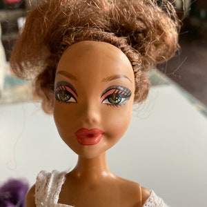以下が含まれることがあります： 茶色の肌をした人形で、茶色の髪をまとめたお団子ヘアスタイルをしています。白いレースのトリムが付いた白いドレスを着ており、ピンクと緑のアイシャドウを施した青い目をしています。