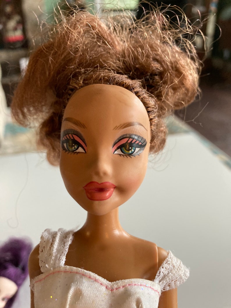 以下が含まれることがあります： 茶色の肌をした人形で、茶色の髪をまとめたお団子ヘアスタイルをしています。白いレースのトリムが付いた白いドレスを着ており、ピンクと緑のアイシャドウを施した青い目をしています。