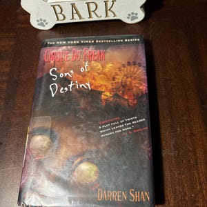 May include: A book titled "Cirque Du Freak: Sons of Destiny" by Darren Shan. The cover features a dark, ominous design with a fiery background and a Ferris wheel. The book is part of "The New York Times Bestselling Series."