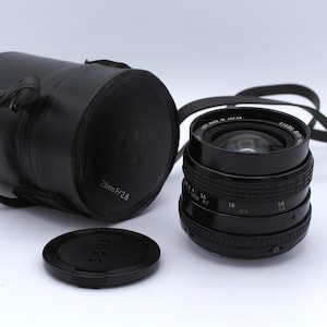 May include: A black Sigma 28mm f/2.8 lens with a black lens cap and a black carrying case. The lens has a green ring with markings for aperture settings.
