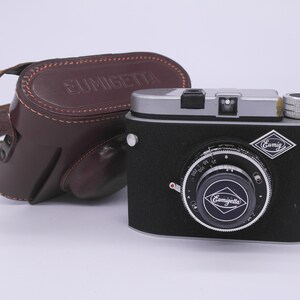 Puede incluir: Una cámara vintage negra y plateada con una funda de cuero. La cámara tiene un objetivo negro con un anillo plateado y el texto "Gumigetta" en él. La funda es de cuero marrón con el texto "Gumigetta" en ella.