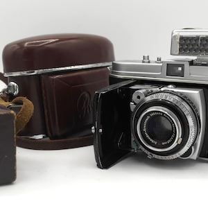 May include: A vintage black and silver 35mm camera with a built-in flash and a leather case. The camera has a lens with a black and silver finish and a silver lens cap. The camera is sitting on a white surface.