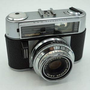 May include: A silver and black Voigtländer Vitomatic II CS camera with a Color-Skopar 2.8/50 lens. The camera has a built-in flash and a rangefinder.