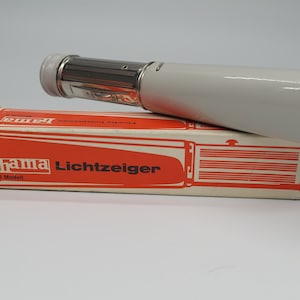 Op de afbeelding: Een witte en zilveren vintage Hama Lichteiger lichtmeter, rustend op de originele oranje en witte doos. De doos heeft het Hama-logo en Duitse tekst. De lichtmeter is cilindrisch.