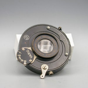 May include: A black camera lens with a silver aperture ring and a silver knob. The lens is labeled "Xenotar Anastigmat 1:2.8/50mm" and "Vario".
