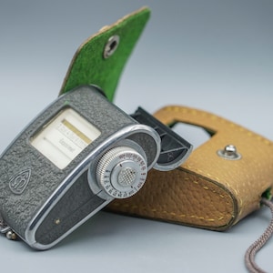 Puede incluir: Un fotómetro vintage gris y plateado con una funda de cuero verde. El fotómetro tiene un dial con números y una aguja. La funda tiene una solapa que se abre para revelar el fotómetro.