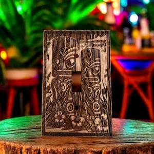 Pode incluir: Uma tampa de interruptor de luz de madeira com um design tiki esculpido. A tampa é de cor castanho escuro com um design esculpido de cor castanho claro. As faces tiki são esculpidas em um estilo tradicional com olhos e bocas grandes.