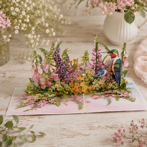 May include: A three-dimensional pop-up greeting card featuring a vibrant garden scene with tall pink and purple foxgloves, yellow wildflowers, and small white blossoms. A large hummingbird with a red throat and a smaller blue bird are perched.