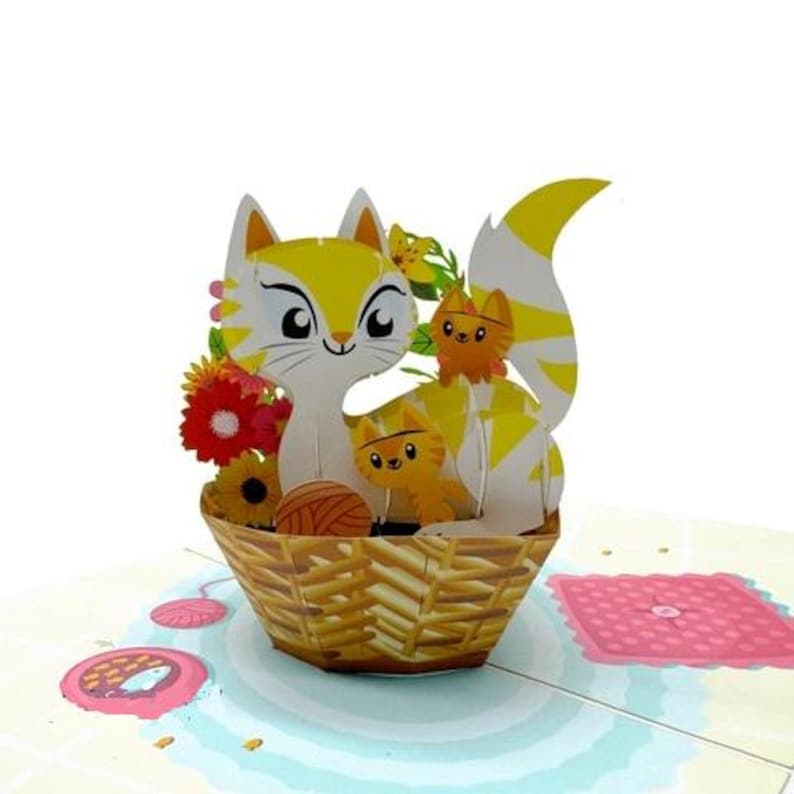 Puede incluir: Una tarjeta de felicitaci&oacute;n emergente en 3D con una familia de gatos en una cesta. El gato es amarillo y blanco con una cola a rayas, rodeado de flores e hilo. La cesta es marr&oacute;n y la tarjeta tiene un fondo azul claro y rosa.
