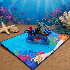 Könnte beinhalten: Pop-up-Grußkarte mit einer Unterwasserszene mit einem Taucher, einem Delfin und bunten Korallen. Die Karte ist blau mit einem dunkelblauen Rand. Ein Seestern und eine Muschel liegen auf der Holzoberfläche.