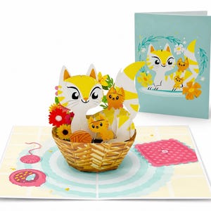 Puede incluir: Tarjeta de felicitaci&oacute;n desplegable con una familia de gatos amarillos y blancos en una cesta tejida con flores. La tarjeta est&aacute; abierta, revelando un dise&ntilde;o 3D, con una ilustraci&oacute;n a juego en la tarjeta cerrada. Tambi&eacute;n se ve un cuadrado rosa y lana.