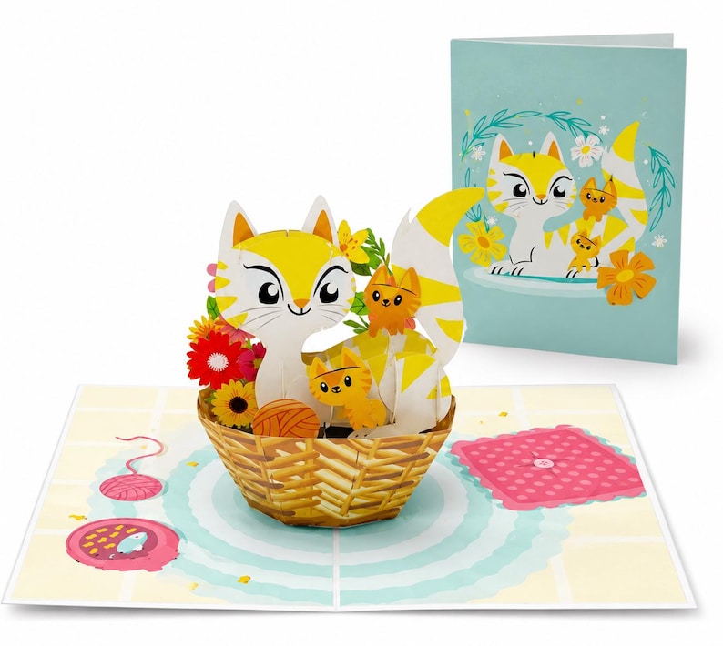 Puede incluir: Tarjeta de felicitaci&oacute;n desplegable con una familia de gatos amarillos y blancos en una cesta tejida con flores. La tarjeta est&aacute; abierta, revelando un dise&ntilde;o 3D, con una ilustraci&oacute;n a juego en la tarjeta cerrada. Tambi&eacute;n se ve un cuadrado rosa y lana.