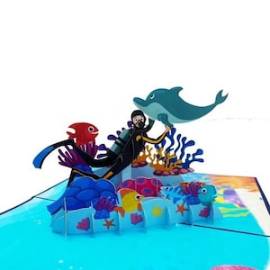 May include: A pop-up card depicting an underwater scene with a scuba diver, a dolphin, and marine life. The card features blue, yellow, red, and orange colours, creating a vibrant and playful design. The card is designed to stand up.