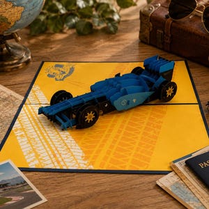 Pode incluir: Cartão pop-up 3D azul e preto de um carro de corrida sobre um fundo amarelo com padrões de pegadas de pneus. O cartão é rodeado por artigos com tema de viagem, incluindo um globo, uma câmara vintage e um passaporte.