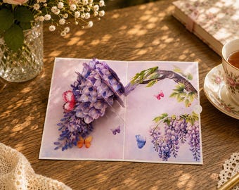 Peut inclure: Carte de vœux pop-up avec un motif de glycine en 3D, présentant des fleurs violettes et des papillons. La carte est ouverte, révélant l'arrangement floral détaillé. Le fond est rose clair, sur une surface en bois.